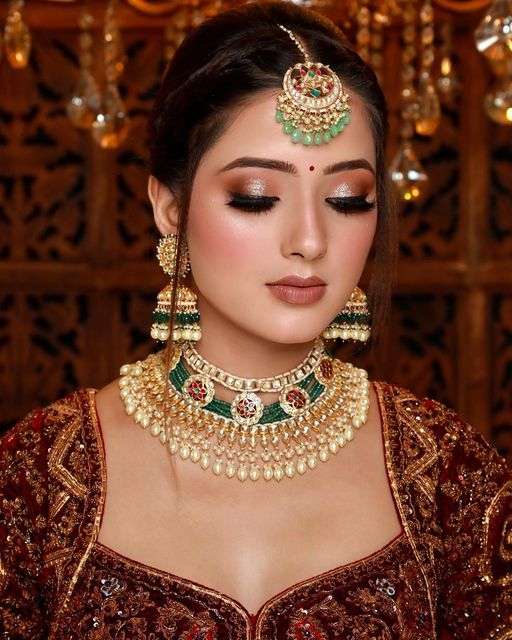 Bride with traditional bridal makeup, elegant eye shadow, nude lips, and intricate jewelry including maang tikka, jhumkas, and layered necklace, wearing a richly embroidered outfit.
