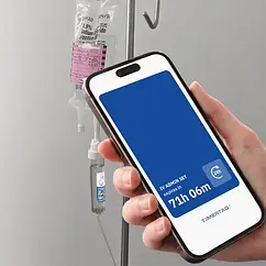 Timertag Pro label affixed to the drip chamber of an IV administration set with a user holding a smartphone near the label. A user experience screen is displayed showing a blue screen with the remaining dwell time for the IV administration set. The dwell time is displayed within a blue screen because the remaining dwell time is more than 2 hours.