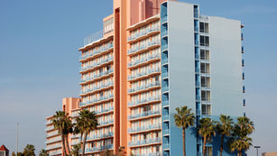 Tall pastel-colored hotel building with balconies, palm trees in front, and a clear blue sky in the background.