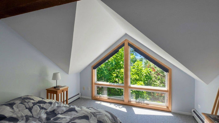An attic bedroom featuring unique trapezoid specialty shape windows left uncovered to look out onto vibrant green trees — BCC of Denver Colorado.; Professional pet-friendly blind designs available from Blind Corners & Curves in Aurora, Steamboat Springs, and Estes Park.; 