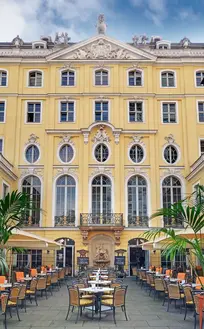 Ehrenhof im Coselpalais Dresden: symmetrische gelbe Barockfassade mit hohen Rundbogenfenstern, Balkon und Wandbrunnen; davor bestuhlte Außenterrasse mit Palmen.