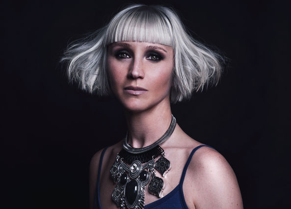 Woman with short platinum blonde hair, bangs, wearing a statement necklace against a dark background.
