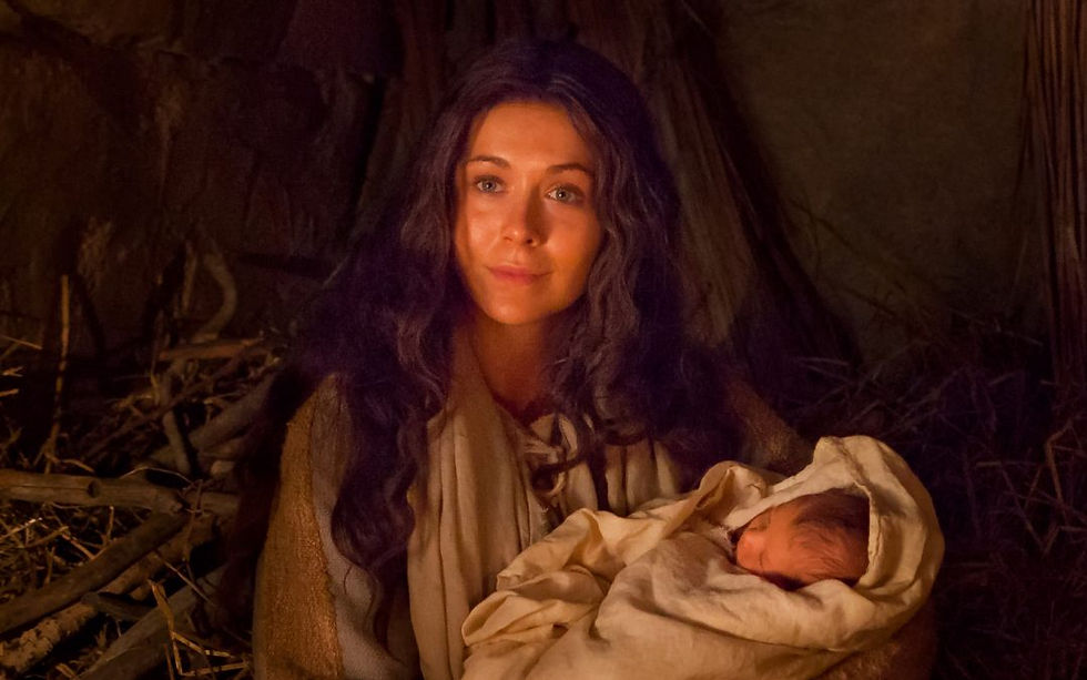 A woman in a rustic setting holds a swaddled baby. Warm lighting creates a serene and tender atmosphere. Hay and wooden elements are visible.
