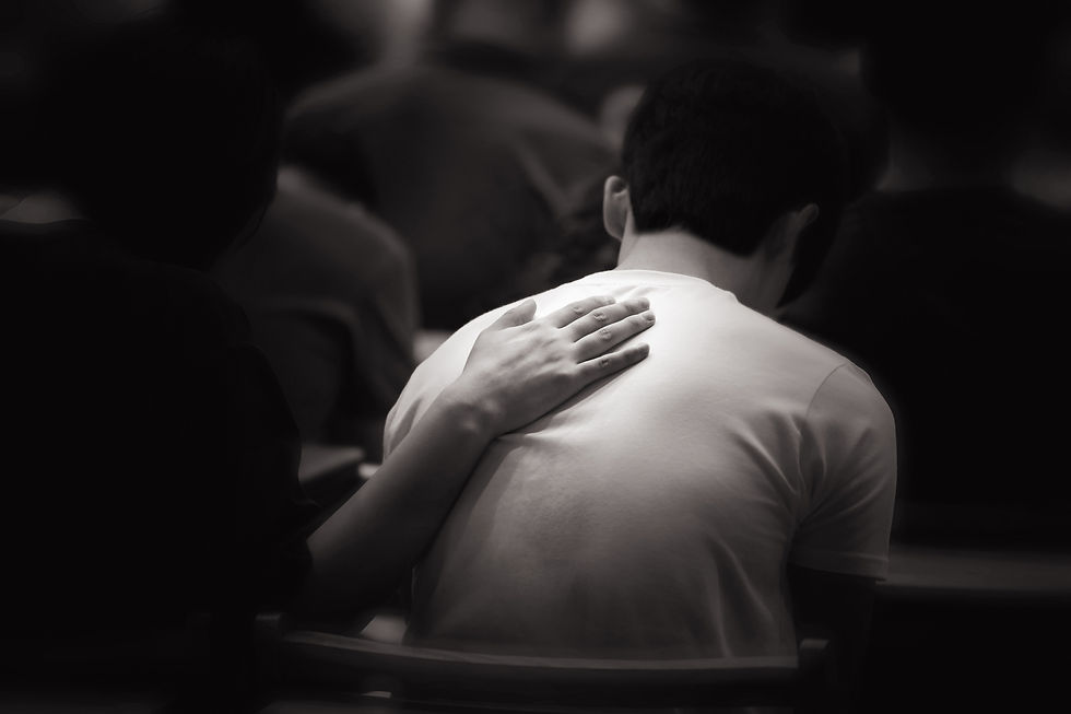 A person in a white shirt is comforted by a hand on their back in a dark room. The mood is supportive and intimate.