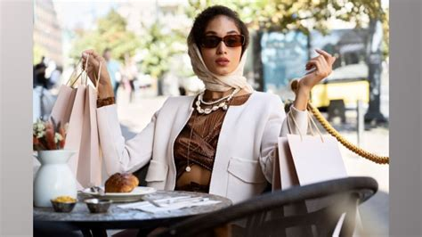 Woman with sunglasses and headscarf sits at outdoor café, holding shopping bags, looking stylish. Pastry on table. Bright, sunny day.