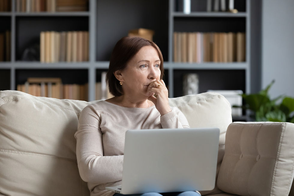 Woman sits on beige sofa with laptop, looking thoughtful. Bookshelf in the background. Bright room with soft lighting, calm mood.
