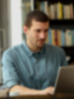 Man in blue shirt focuses on a laptop in a cozy room with a bookshelf in the background. Warm lighting, relaxed mood.