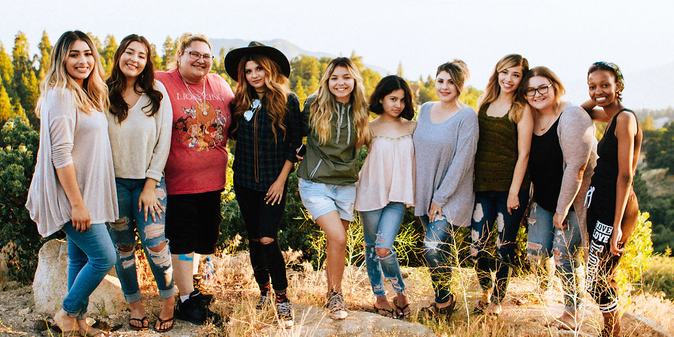 A group of nine women smiling outdoors in a sunny, forested area. They wear casual clothes; one has a hat and another a Lion King shirt.