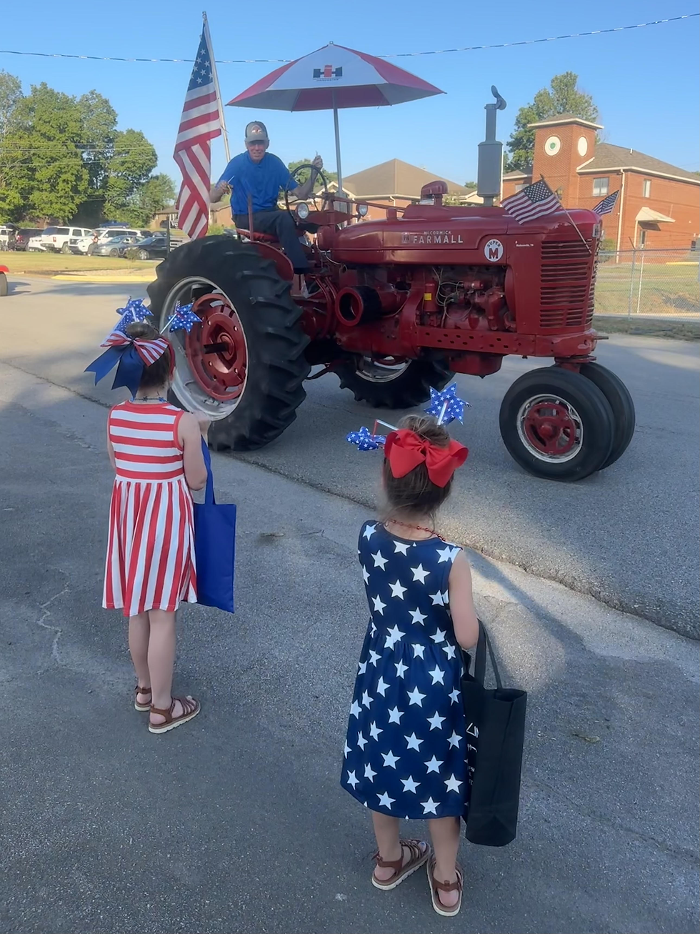 Two little girls in patriotic costumes watch a man on a tractor drive by
