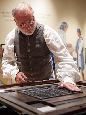 Photo of Charlie Rhodarmer, a man with a white beard, wearing a 18th century costume, works a printing press in a museum.