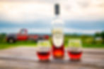 A bottle of muscadine wine with two glass cups beside it. Outside on a wooden table with a truck and mountains in the background.