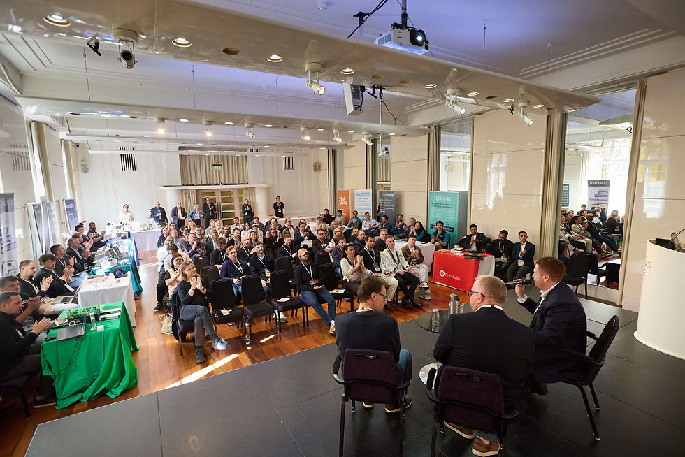 Host Planet Roadshow London. A conference room with three speakers on stage facing an audience. Attendees sit at tables with green and red cloths, applauding. Bright lighting.