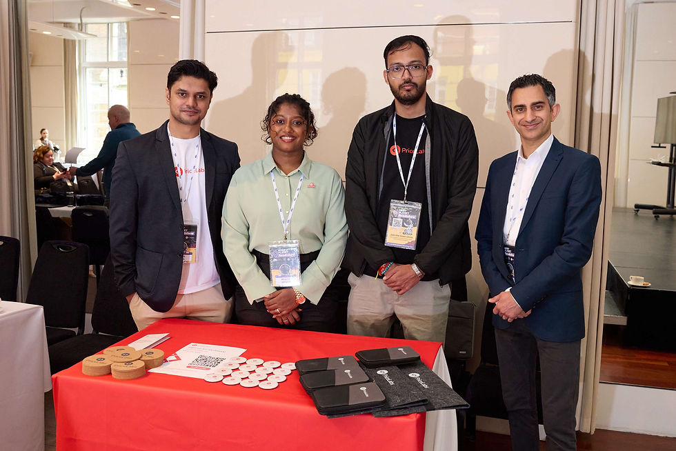 PriceLabs staff attend Host Planet Roadshow London. Four people stand smiling behind a table with promotional items. They wear name tags. Background shows a bright conference setting.