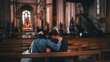 A young couple sits closely together on a wooden church pew, embracing each other in the peaceful interior of a grand historic church. Warm light illuminates the ornate altar, towering columns, and carved wooden pulpit, creating an intimate contrast between human connection and the impressive sacred architecture.