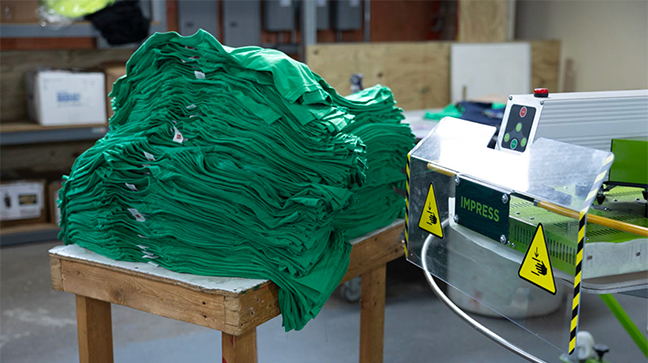 ROQ IMPRESS Automatic Transfer Machine next to a stack of green shirts