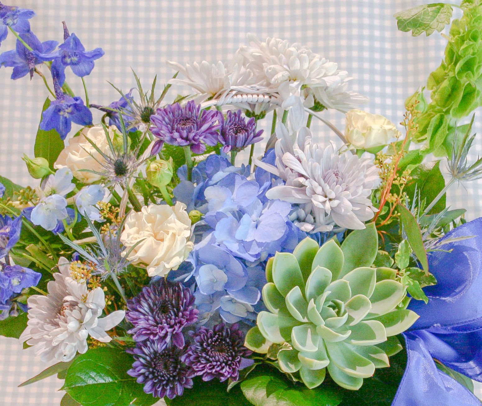 A close up a floral design full of blue, white and green flowers like hydrangea, roses, and mum all designed within a basket.