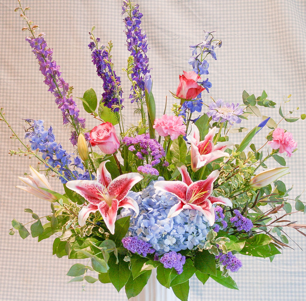 a fresh floral arrangement featuring stargazer lilies, hydrangea, roses, irises, delphinium, larkspur in a tall white vase