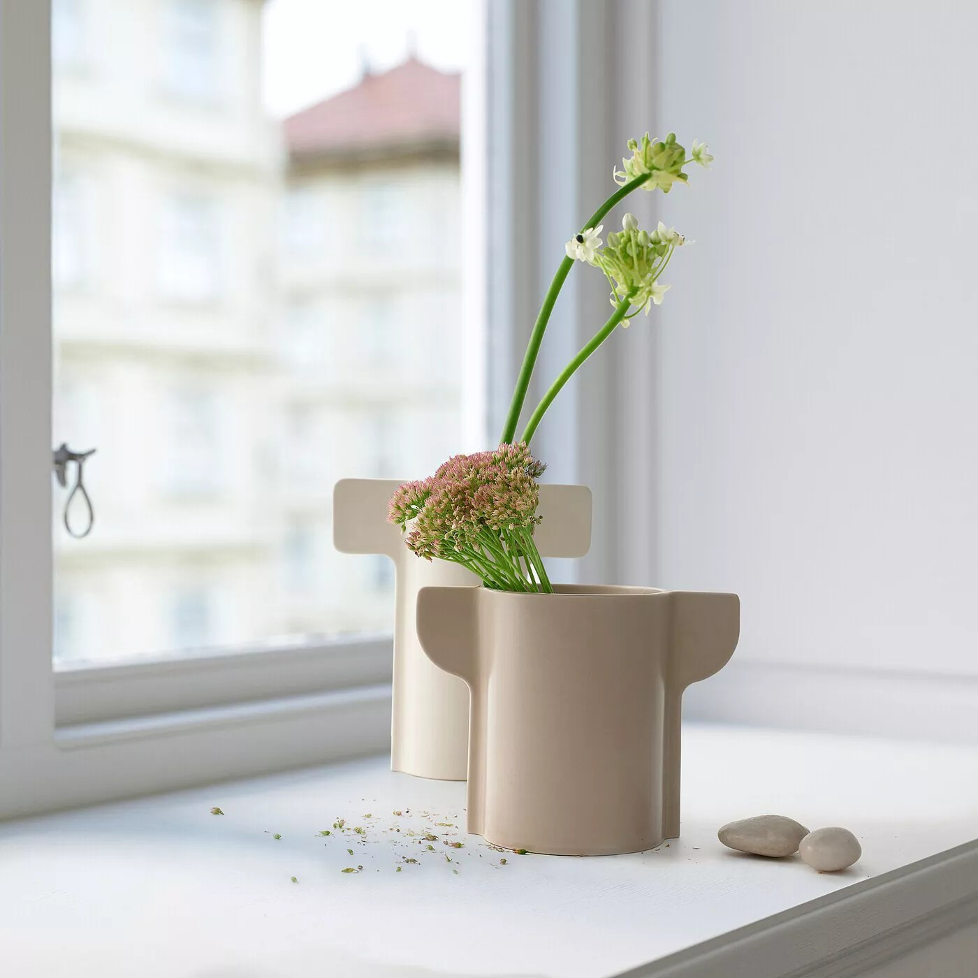 Pelarönn vase by designer Gustav Carlberg—a sculptural beige vase with two distinctive handles, placed on a white windowsill. It holds a delicate arrangement of green and pink flowers, with scattered petals and pebbles adding texture to the serene composition. Soft natural light filters through the window, casting a gentle glow on the scene.