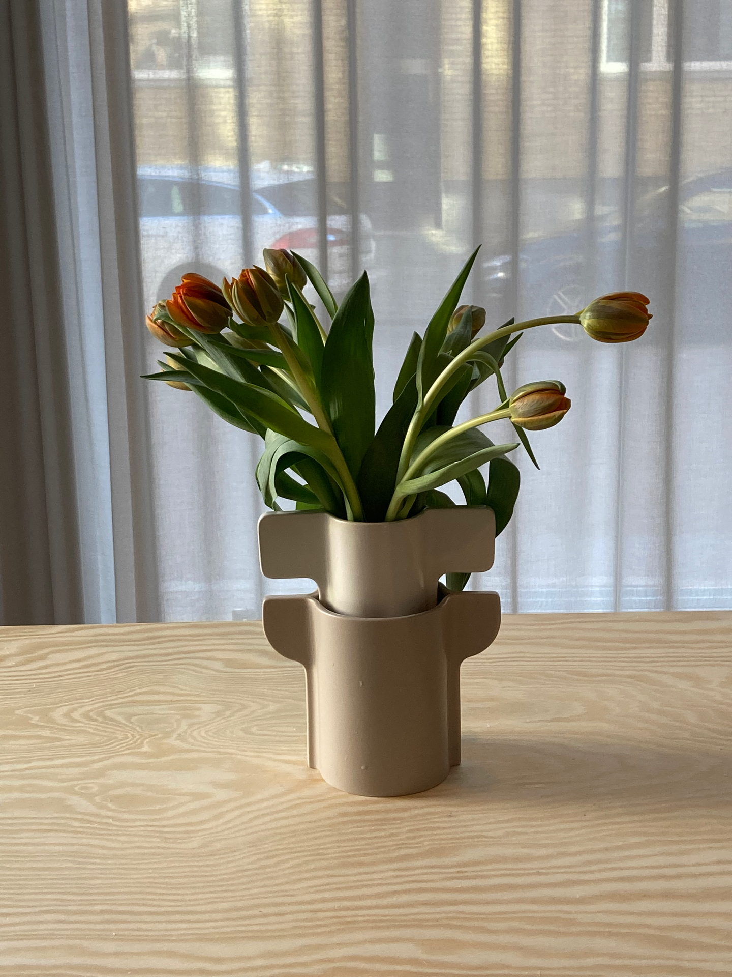 Pelarönn vase by designer Gustav Carlberg—a sculptural beige vase with distinctive handles, holding a bouquet of orange tulips. Placed on a wooden table near sheer curtains, the soft lighting enhances the elegant and natural aesthetic.