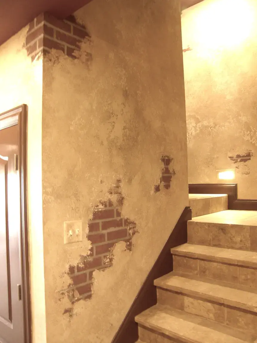 Warm-lit staircase hallway with faux plaster walls peeling to reveal brick patches and tan tile steps