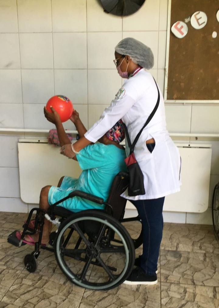 Profissional da saúde atendendo idosa em cadeira de rodas realizando exercício com bola no abrigo bom jesus