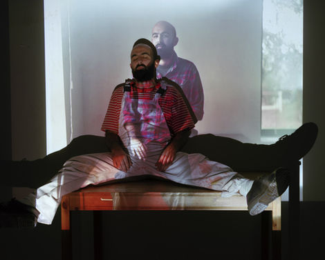 דמות גבר יושב בפישוק רגליים על ספסל נמוך, עליו מוקרנת דמות נוספת. נוצר שילוב בין גוף נוכח לבין היעדר — צללים של תודעה, זיכרון או נשמה. תחושת כפילות או זהות מתפצלת.