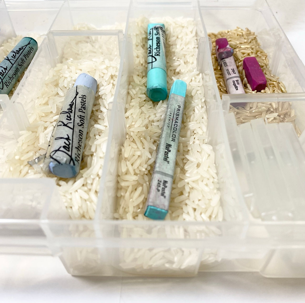 Chalk pastels in a clear container, shot from above looking down. There is rice underneath the pastels.