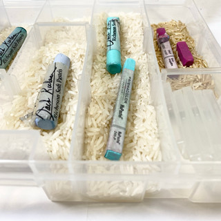 Chalk pastels in a clear container, shot from above looking down. There is rice underneath the pastels.