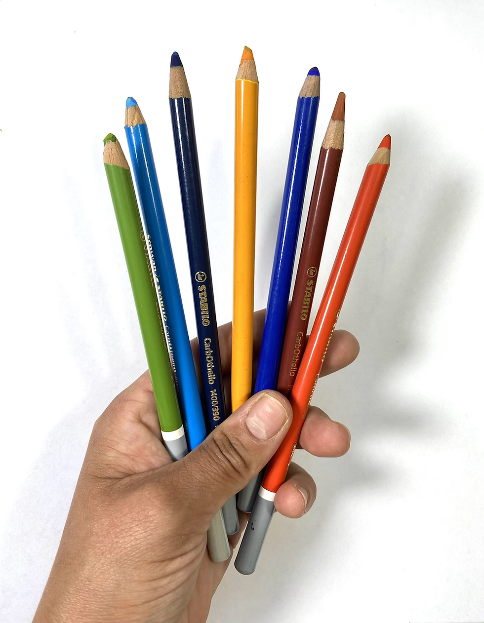 A hand is holding chalk pastel pencils, they are fanned out to show the different colours. The background is minimal and light.