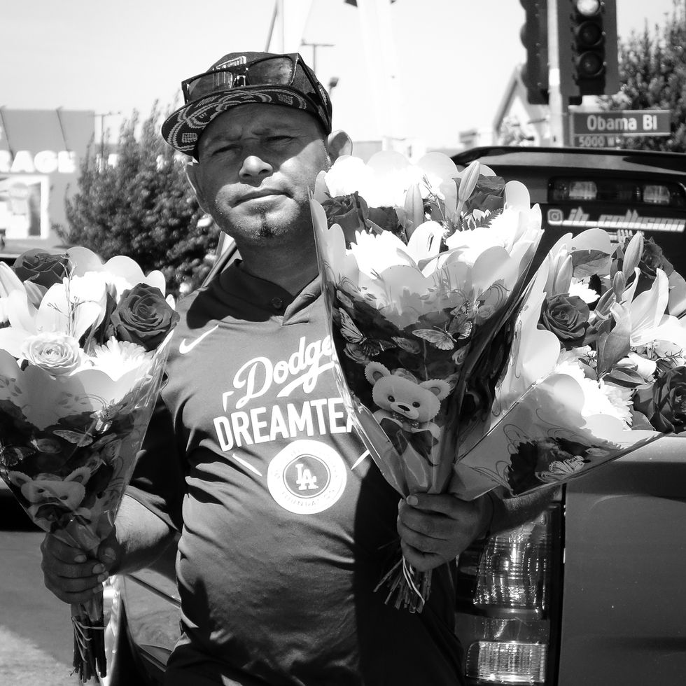 A fine art print of a man wearing a baseball cap and a Dodgers shirt holding several floral arrangements while standing next to a parked truck near an intersection marked with a traffic signal. Captured by photographer Alexander Eastick in California.