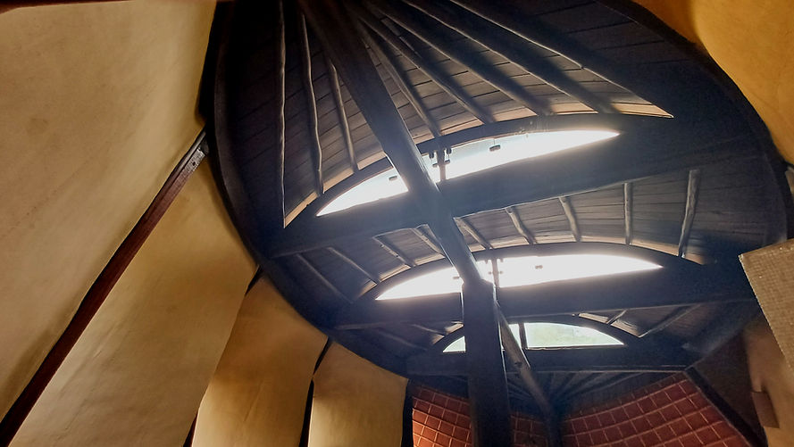 Claraboias em formato de meia-lua filtrando a luz zenital sob estrutura de madeira roliça e paredes de terra de arquitetura orgânica.