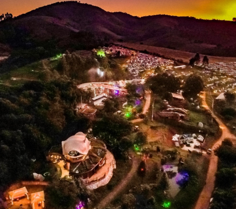 Vista aérea noturna de habitação ecológica circular com telhado em espiral iluminada por luzes quentes e caminhos de LED na floresta. Design biofílico e arquitetura sustentável por César Augusto Arquiteto.