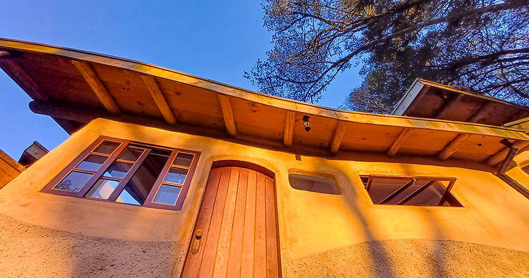 Fachada Oeste e entrada com porta em arco de madeira natural integrada à parede com elementos curvos entre reboco de terra e barra de proteção inferior e curvatura delicada da cobertura, na Casa Nave Terra, com foco em bioconstrução.