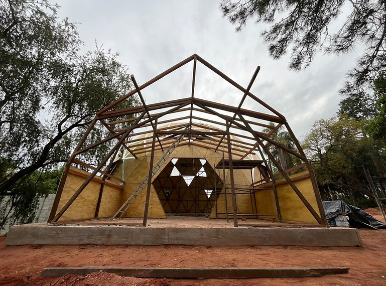 Estrutura em madeira do pórtico da antesala de domo geodésico em fase de construção sobre fundação em chácara arborizada — design de geometria e bioconstrução limpa por César Augusto Arquiteto Ecológico