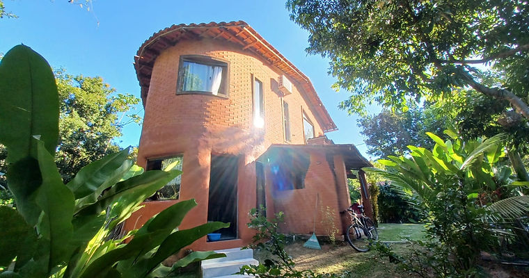 Fachada curva da Casa Luz do Arraial com reboco de terra areia e tijolos de solo-cimento, integrada ao paisagismo tropical em projeto de bioconstrução.
, construção sustentável de verdade.