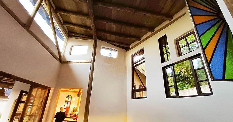 Sala com pé-direito duplo em arquitetura sustentável exibindo pilares de troncos de madeira natural, paredes de terra com reboco natural e tinta de cal, detalhe de vitral colorido.