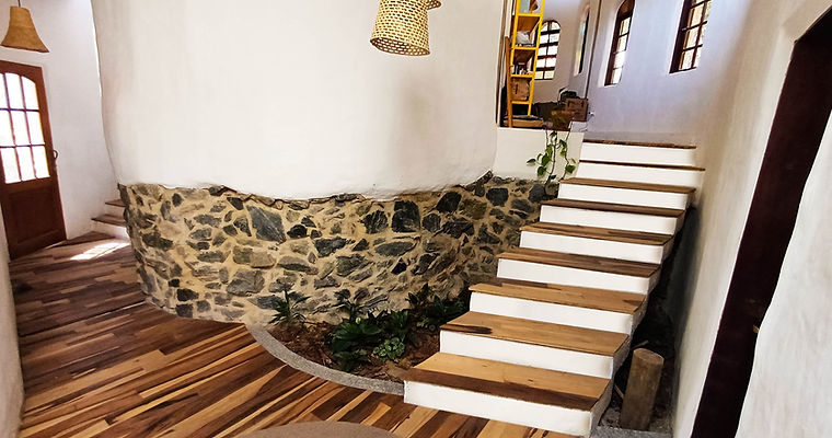 Hall interno da Casa Nave Terra exibindo parede curva de reboco natural sobre base de pedra bruta, piso de madeira e canteiro integrado em construção saudável e biofília.