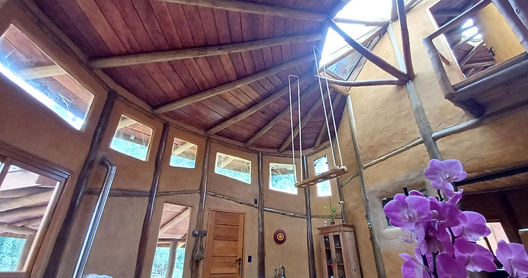 Interior de casa de terra com pé-direito duplo, teto de vigas radiais de madeira e luminária balanço suspensa. Projeto de arquitetura sustentável assinado por César Augusto Arquiteto.