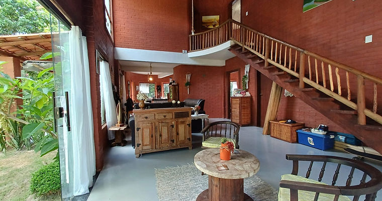 Interior da sala de estar da Casa Luz do Arraial com paredes de tijolos ecológicos aparentes, piso de cimento queimado e escada de madeira, focando em construção saudável.