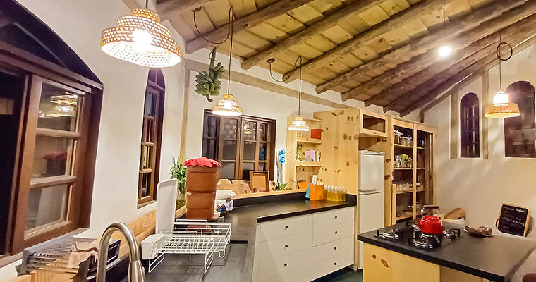 Cozinha rústica planejada em casa de bioconstrução com vigamento de madeira roliça, paredes curvas de reboco natural e luminárias de palha.