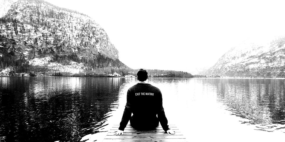 Person sits on dock, observing serene lake and mountains. Sweater reads "EXIT THE MATRIX." Calm, reflective mood.
