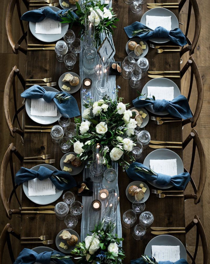 Elegant table setting with white roses, candles, blue napkins, and menus on plates. Wooden table, rustic chairs, cozy ambiance.