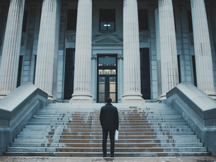 A person stands at the bottom of large courthouse steps holding paperwork, facing the entrance of a government building.