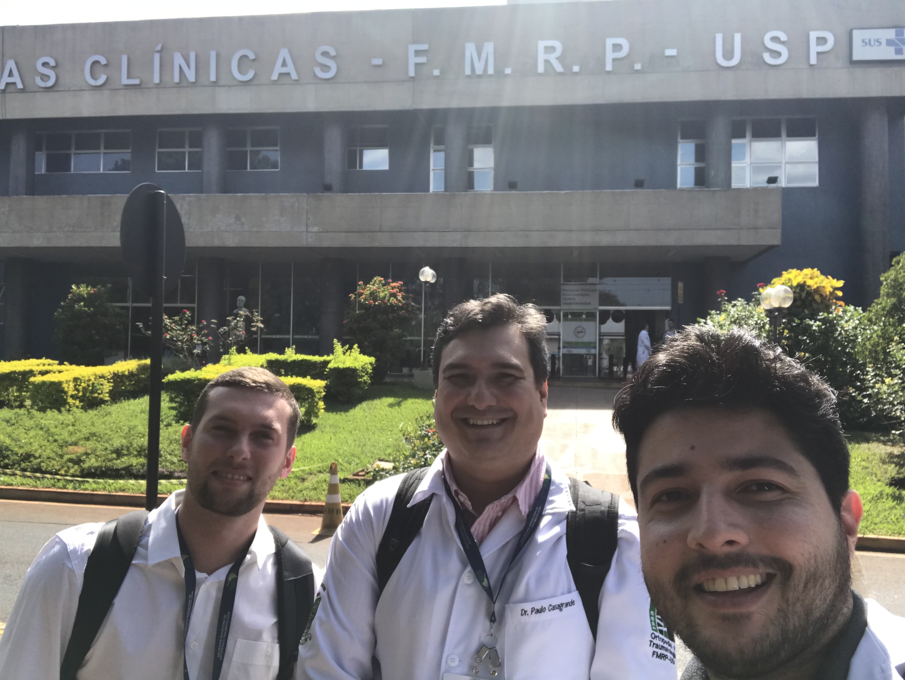 Três médicos sorrindo na frente de um hospital.