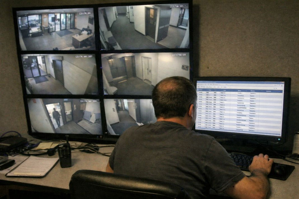 Person monitoring multiple security camera screens and a computer, displaying surveillance footage and data in a dimly lit control room.