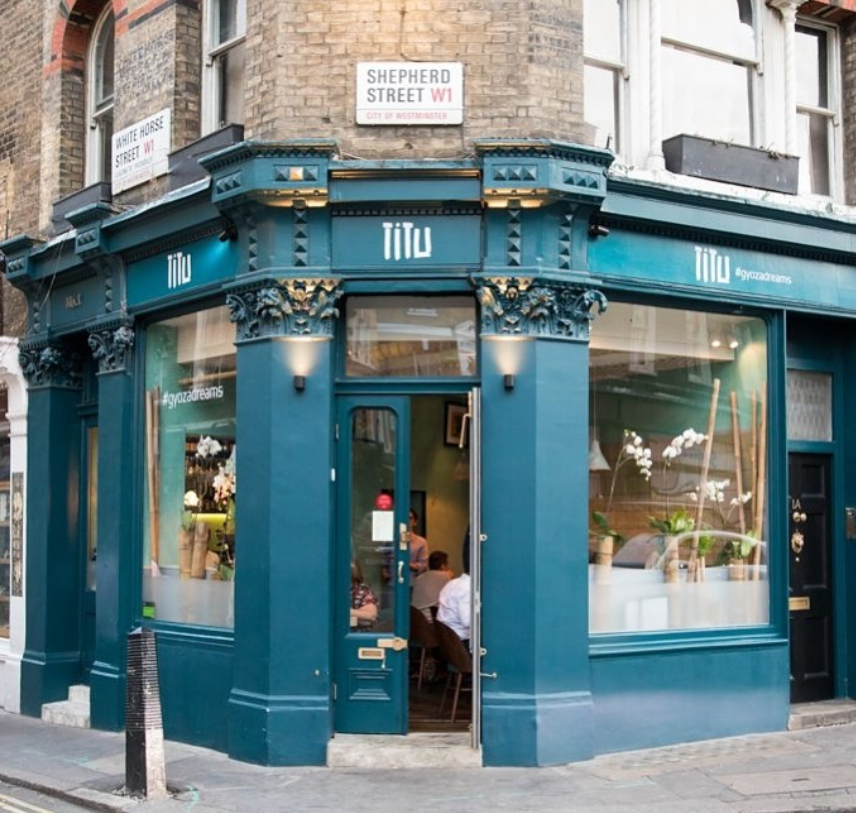 Blue restaurant corner with ornate details, "Titu" sign, and visible diners inside. Street signs read "Shepherd St W1" and "#gyozadreams."