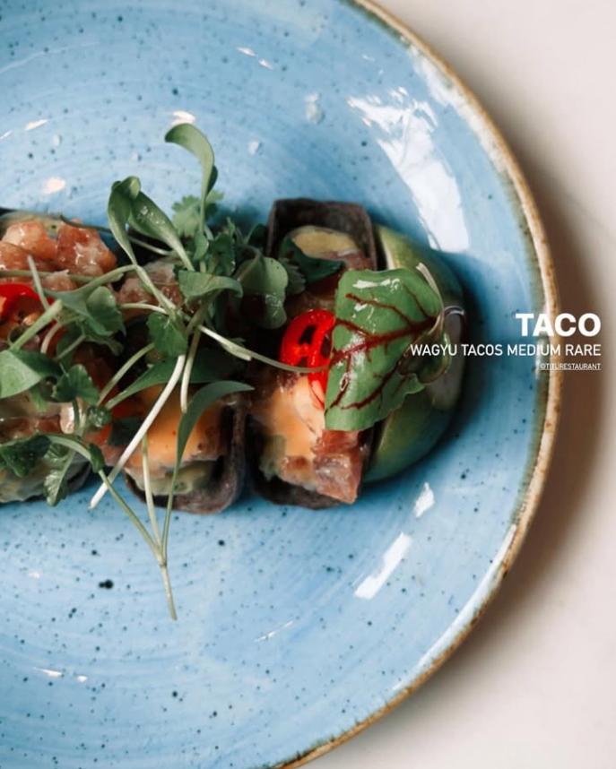 Wagyu tacos on a blue plate with greens and red chili, garnished artfully. Text reads "Taco Wagyu Tacos Medium Rare." Mood is fresh and vibrant.