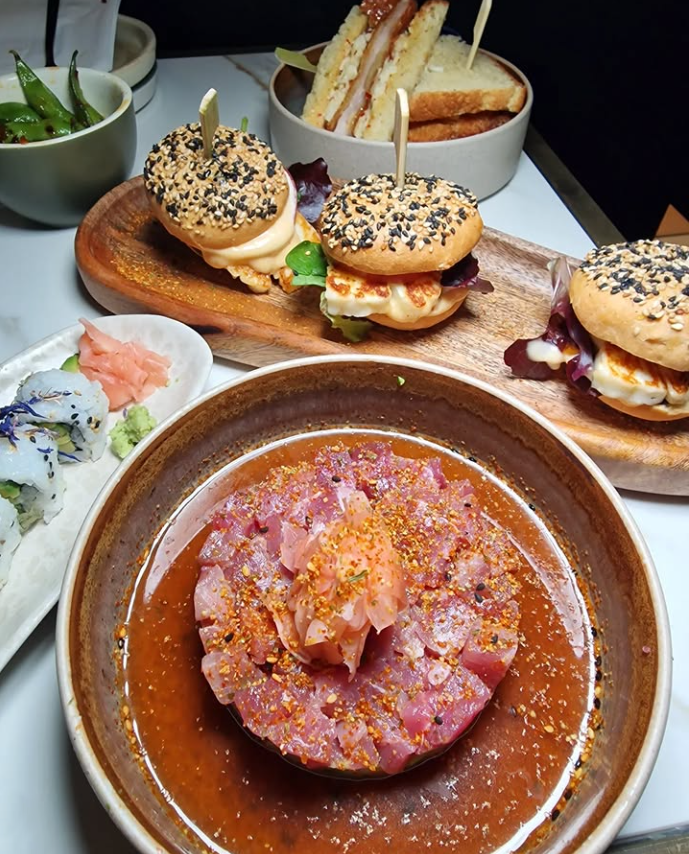 Assorted food on a table: sushi, sliders with seeded buns, a sandwich, and a bowl of pink tartare with seasoning, creating a gourmet spread.