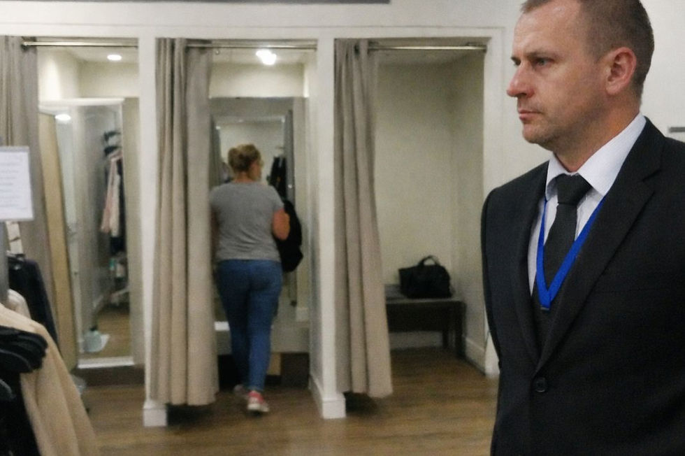 A woman enters a changing room in a clothing store, holding a bag. A man in a suit watches, looking serious. Gray curtains and wooden floor.