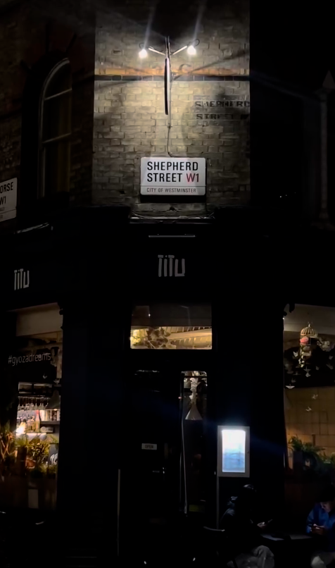 Street scene at night with brick building lit by streetlights. Shepherd Street W1 sign visible. People on sidewalk near a restaurant.